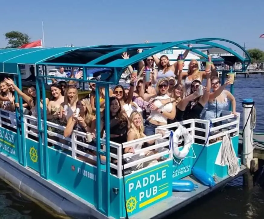 A boatful of guests waving from the rail of a branded Paddle Pub boat