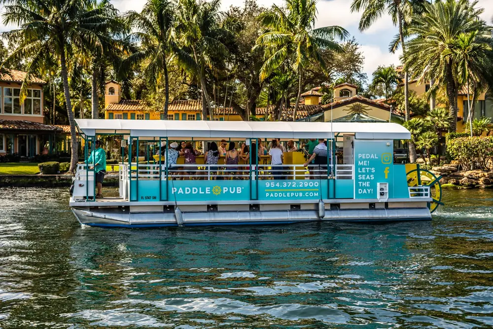 A branded Paddle Pub boat cruising a palm-lined waterway, guests gathered along the bar rail